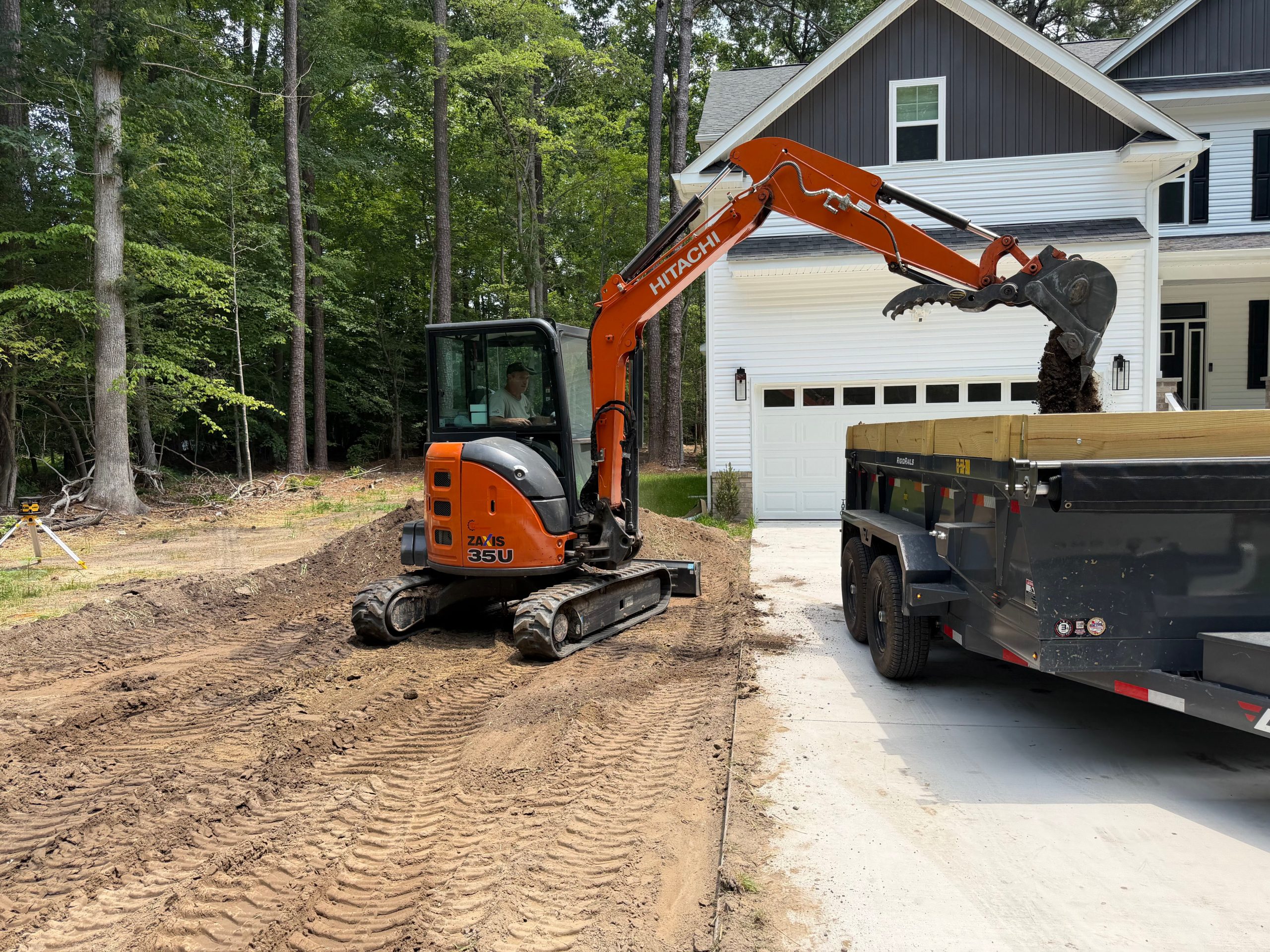 Excavator working in a driveway next to a house garage with a dump trailer attached